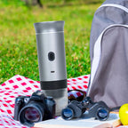 A portable silver coffee maker with a black grip stands on a red-and-white checkered blanket outdoors on grass, alongside a gray backpack, camera, binoculars, sunglasses, and an open book.